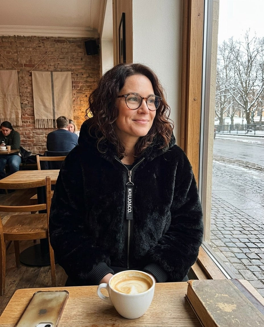 Stina, fuskpälsjacka med huva för dam i svart, stylinginspiration på ett kafé