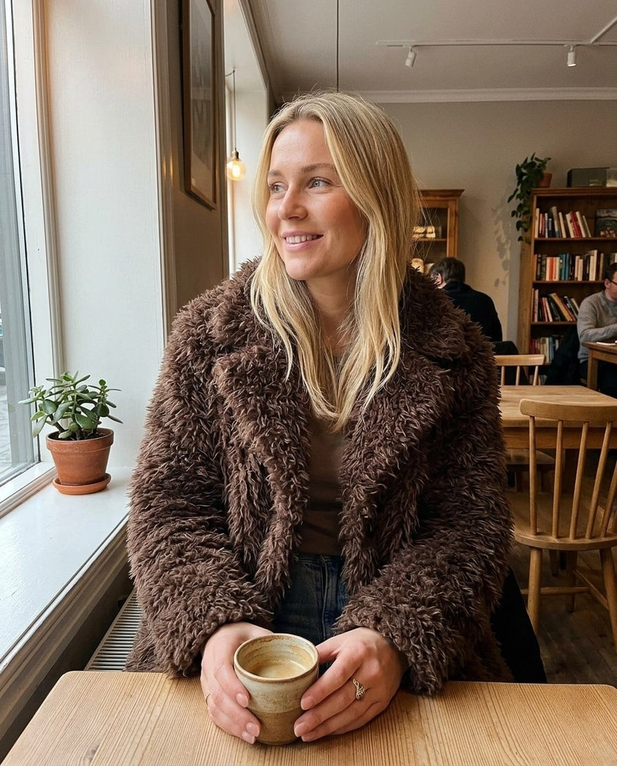 Saga, lång fuskpälskappa för dam i brun, stylinginspiration på ett kafé