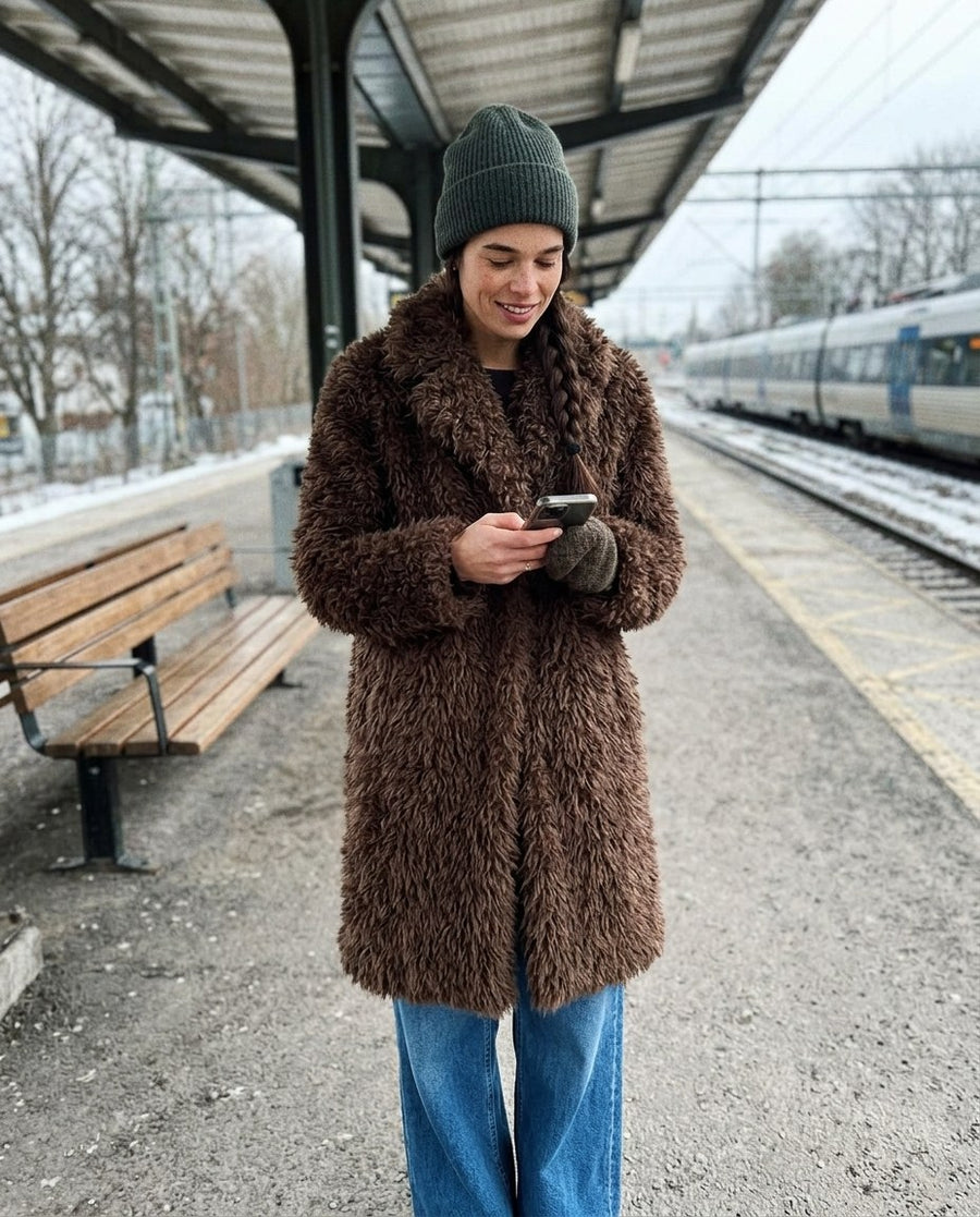 Saga, lång fuskpälskappa för dam i brun, stylinginspiration på en perron
