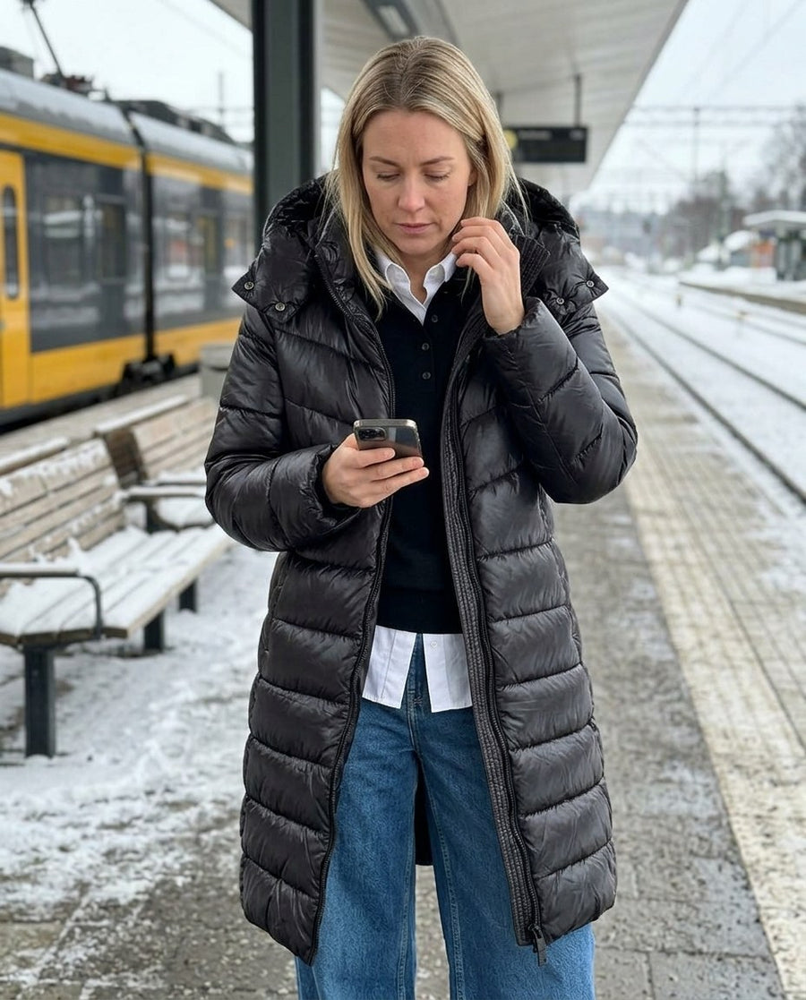 Vera, lång pufferkappa med huva för dam i svart, stylinginspiration på en perron
